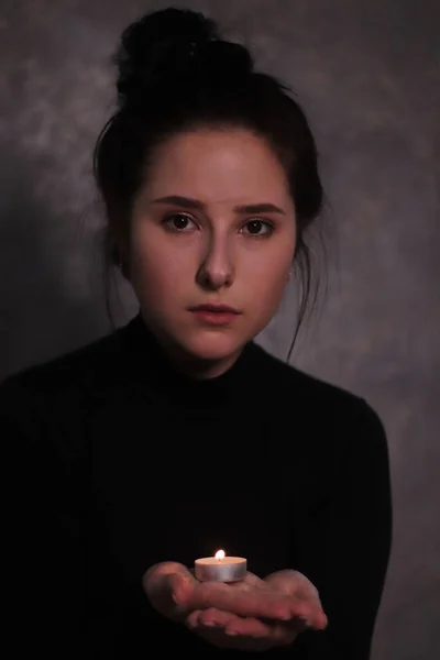 Oscuro retrato de una chica morena triste casi estalló en lágrimas en un cuello alto negro sosteniendo vela contra una pared gris — Foto de Stock