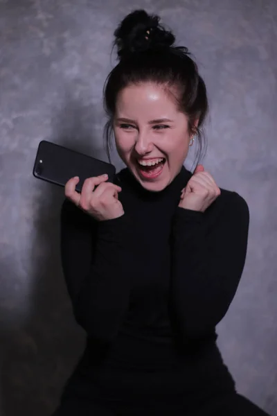 Érzelmi portré egy boldog barna lány egy fekete garbó tartó fekete telefon egy szürke fal — Stock Fotó