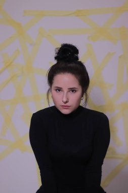 Siyah balıkçı yaka kazaklı esmer bir kızın portresi. Beyaz bir duvarda sarı çizgili bir arka plan.