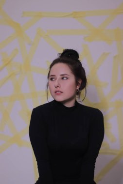 Siyah balıkçı yaka kazaklı esmer bir kızın portresi. Beyaz bir duvarda sarı çizgili bir arka plan.