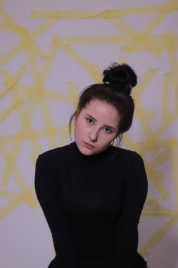 Siyah balıkçı yaka kazaklı esmer bir kızın portresi. Beyaz bir duvarda sarı çizgili bir arka plan.