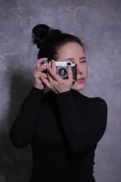 Retrato de una chica morena en un cuello alto negro sosteniendo una vieja cámara retro contra una pared gris — Foto de Stock