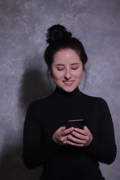 Emotioneel portret van een brunette meisje in een zwarte coltrui met zwarte telefoon tegen een grijze muur — Stockfoto