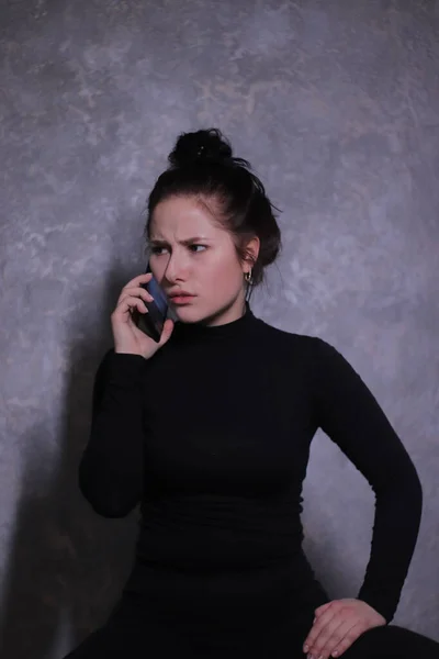 Συναισθηματικό πορτραίτο μιας μελαχρινής κοπέλας με μαύρο ζιβάγκο που αγανάκτησε μιλώντας στο τηλέφωνο σε έναν γκρίζο τοίχο — Φωτογραφία Αρχείου
