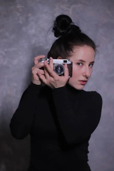 Portrait d'une fille brune au col roulé noir tenant une vieille caméra rétro contre un mur gris — Photo