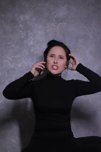 Retrato emocional de uma menina morena em uma gola alta preta falando indignamente ao telefone contra uma parede cinza — Fotografia de Stock