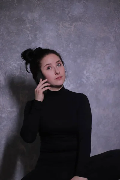 Emotional portrait of a brunette girl in a black turtleneck indignantly talking on the phone against a gray wall — Stock Photo, Image