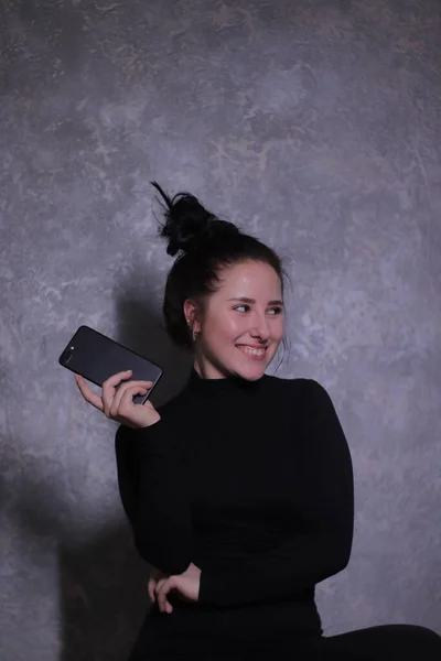 Emotivní portrét šťastné brunetky dívky v černém roláku drží černý telefon na šedé stěně — Stock fotografie