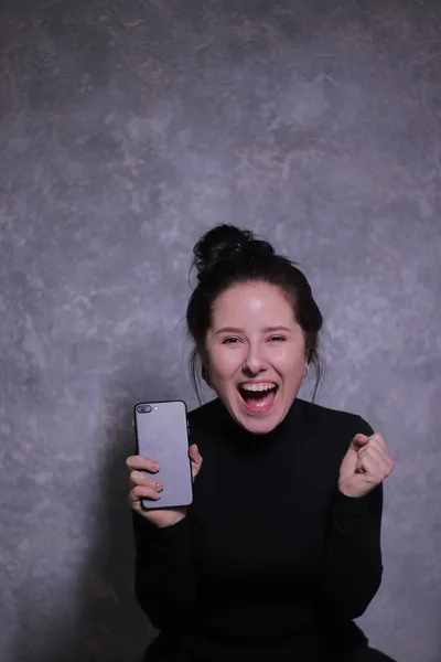 Retrato emocional de una chica morena en un cuello alto negro sosteniendo el teléfono negro contra una pared gris —  Fotos de Stock