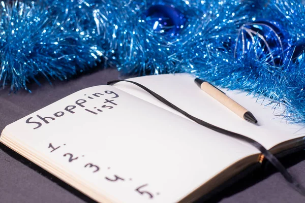 A notebook with title "Shopping list" with a pen near a blue shiny tinsel for christmas tree. new years theme — Stock Photo, Image