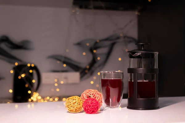 French press and tea in double-bottom glass next to braided balls for decor on a background of golden yellow bokeh — Stock Photo, Image