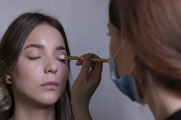 Prozess des Schminkens einer schönen Brünetten durch einen Meister in einer medizinischen Maske. Schönheits-, Modellierungs- und Kosmetikkonzept — Stockfoto