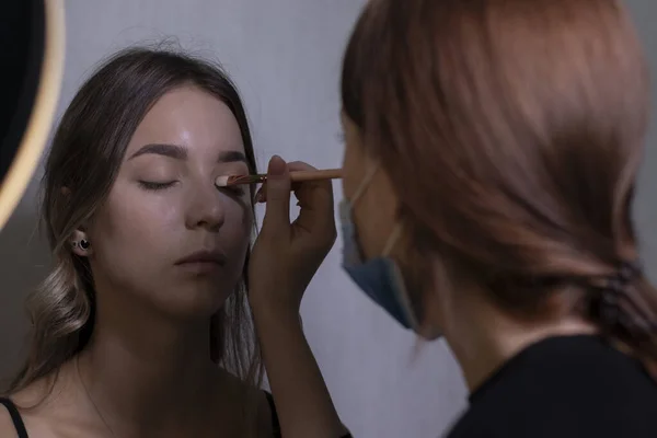 Proceso de aplicación de maquillaje a una hermosa morena por un maestro en una máscara médica. concepto de belleza, modelado y cosmética —  Fotos de Stock