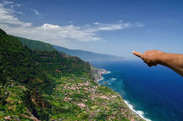 Hand of male tourist pointing at view of north coast of Madeira island, Santana town surrounded by high mountains and deep blue Atlantic ocean. Popular tourist destination in Madeira, Portugal. Space for text.