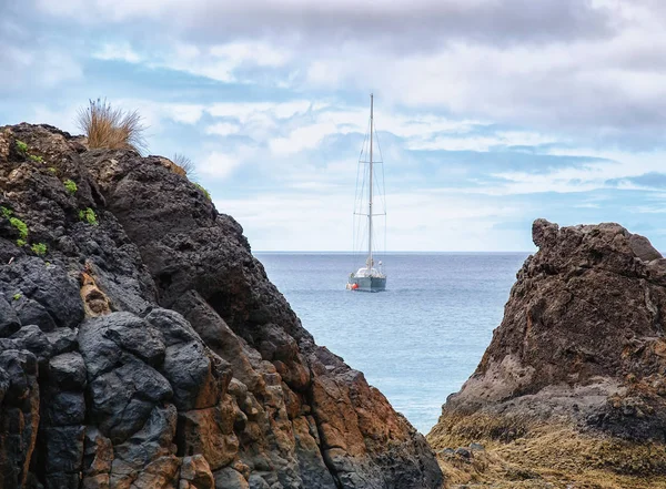 Elegant yacht is sailing towards the coastline of Madeira island. Colorful natural volcanic rocks with few plants at the foreground. Summer vacation concept.
