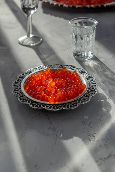 Natural red caviar on silver plate on gray background. Vodka shots and plate of caviar at unfocused background. Selective focus. Healthy eating concept. Russian style food background with copy space.
