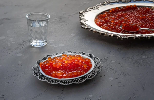 Natural red caviar on silver plate on gray background. Vodka shots and plate of caviar at unfocused background. Selective focus. Healthy eating concept. Russian style food background with copy space.