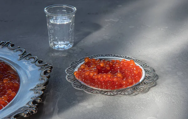 Natural red caviar on silver plates on gray background. Vodka shotr at unfocused background. Selective focus. Healthy eating concept. Russian style food background with copy space. Horizontal orientation.