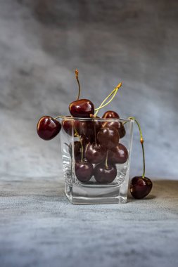 Olgun koyu kırmızı ve camda siyah kirazlar. İki tatlı kiraz sarkmış ve meyve dolu bardaktan düşüyor. Kopyalama alanı olan dikey gri arkaplan. Sağlıklı beslenme ve beslenme kavramı