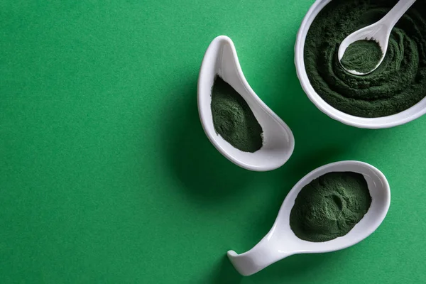 Green chlorella or spirulina powder in three differently shaped white porcelain bowls on green paper background. Healthy superfood eating and dieting concept. Horizontal orientation. Large copy space.