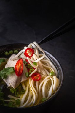 Baharatlı Asya çorbasının yakın çekimi. Tavuk buili ve et, kırmızı biber, kişniş, soğan, Japon eriştesi, yemek çubukları tarafından alınan siyah çorba kasesi. Siyah arka plan. Seçici odaklanma. Boşluğu kopyala.