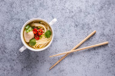 Baharatlı Asya çorbası. Tavuk bulyonu ve et, kırmızı biber, kişniş, soğan ve Japon eriştesi gri yüzeyde tahta çubuklarla birlikte beyaz kasede. Yatay oryantasyon. Uzayı kopyala Düz yatıyordu.