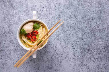 Baharatlı Asya çorbası. Tavuk bulyon ve et, kırmızı biber, kişniş, soğan, gri yüzeyde beyaz kasede Japon eriştesi. Tahta yemek çubukları kasede. Yatay oryantasyon. Uzayı kopyala Düz yatıyordu