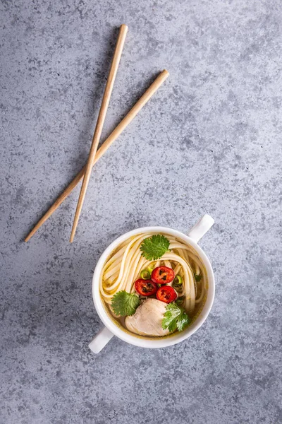 Baharatlı Asya çorbası. Tavuk bulyonu ve et, kırmızı biber, kişniş, soğan ve Japon eriştesi gri yüzeyde tahta çubuklarla birlikte beyaz kasede. Dikey oryantasyon. Uzayı kopyala Düz yatıyordu.