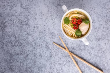 Asya usulü tavuk çorbası. Bouillon, et, kırmızı biber, kişniş, soğan, Japon eriştesi gri masada. Tahta çubuklar kasenin yanından geçti. Yatay oryantasyon. Uzayı kopyala Düz yatıyordu.