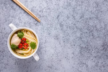 Asya usulü tavuk çorbası. Bouillon ve et, kırmızı biber, kişniş, soğan, gri yüzeyde beyaz kasede Japon eriştesi. Kasenin yanında tahta çubuklar var. Yatay oryantasyon. Uzayı kopyala Düz yatıyordu.