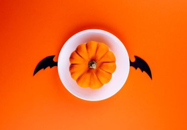 Beyaz yuvarlak tabakta çiğ turuncu balkabağı elle oyulmuş siyah yarasa kanatlarıyla birlikte yatay turuncu kağıt arka planda. Uzayı kopyala Yaratıcı düz yatak. Cadılar Bayramı sembolleriyle komik bir görüntü.