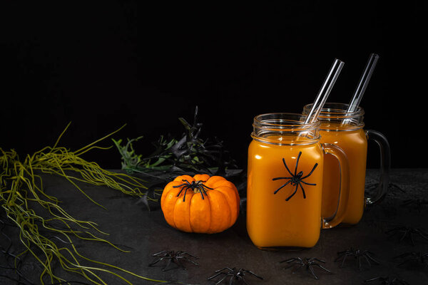 Glass jars with pumpkin mocktail and glass straws on black textured table decorated with spiders, spooky plants and pumpkin. Halloween decor. Horizontal background with copy space. Selective focus.