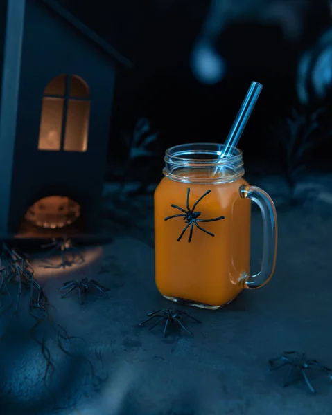 Halloween scary still life with pumpkin mocktail, spiders, spooky black house with light in windows and plants. Strange shadows and silhouettes. Black textured surface. Selective focus.