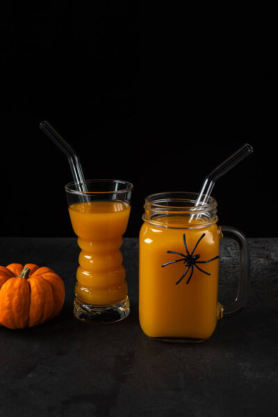 Non-alcoholic pumpkin mocktail in drinking glass and square jar with glass straws and whole orange pumpkin on black textured surface. Halloween spider decor on jar. Healthy eating concept. Copy space.