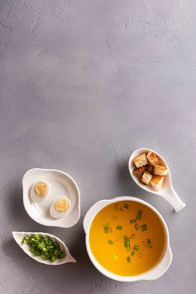 Bouillon in white soup bowl, chopped spring onions or scallions, quail eggs and croutons on gray stone background. Table top view. Vertical orientation. Healthy eating and dieting concept. Copy space. — Stock Image Bouillon White Soup Bowl Chopped Spring Onions Scallions Quail Eggs Stock Picture
