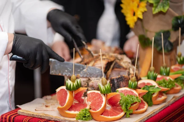 Chief cook cuts into pieces freshly cooked pork hip, process close up. Decorations of fruits and vegetables on the table, catering. Show at the restaurant