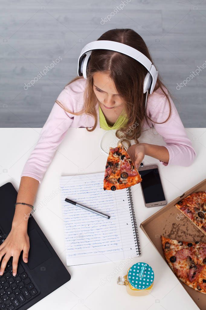 Young teenager girl working on a school project - having a snack over her notebook using her laptop, smartphone and eating simultaneously