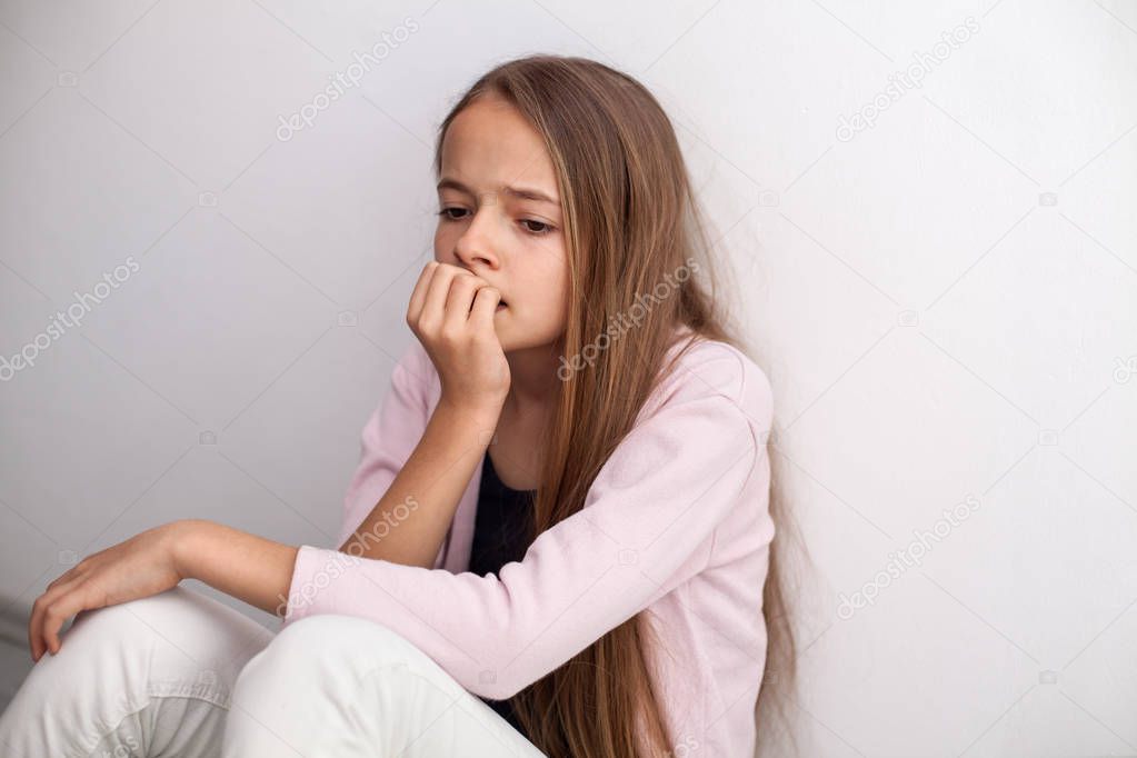 Worried teenage girl biting her nails sitting on the floor by the wall - looking agitated