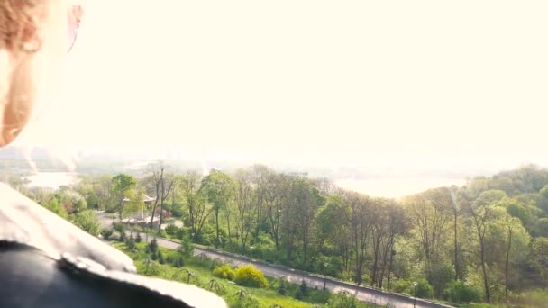 Европейская девушка улыбается в камеру и слушает музыку в солнцезащитных очках на фоне большого города — стоковое видео