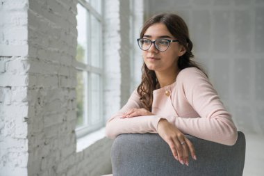 Genç Sevimli seksi sevimli kız elbise ve gevşek saç gri bir koltukta oturan büyük pencereler ile bir beyaz odada bardaklarda gebe