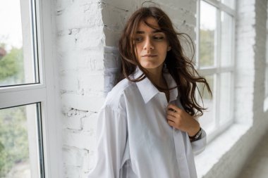 Genç Sevimli seksi sevimli kız elbise ve gevşek saç gri bir koltukta oturan büyük pencereler ile bir beyaz odada bardaklarda gebe