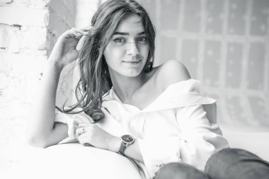 beyaz bir adamın gömleği gevşek bir uzun saçlı parlak bir oda penceresinde yakınındaki beyaz bir kanepede oturan bir genç, çok seksi, sevimli kız portresi 