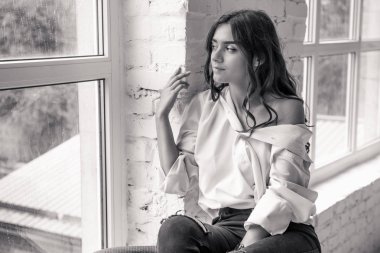 beyaz bir adamın gömleği gevşek bir uzun saçlı parlak bir oda penceresinde yakınındaki beyaz bir kanepede oturan bir genç, çok seksi, sevimli kız portresi 