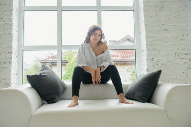 Genç Sevimli seksi sevimli Gözlüklü kız çıplak sarıl bağsız kıl ile büyük bir pencere yakınında ile beyaz bir erkek ceket beyaz kanepe oturur gebe