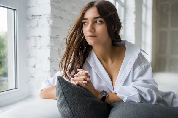 young pretty sexy charming girl in a white man\'s shirt sits on a white sofa near the window in a bright room with a long hair lying around where the glasses lie