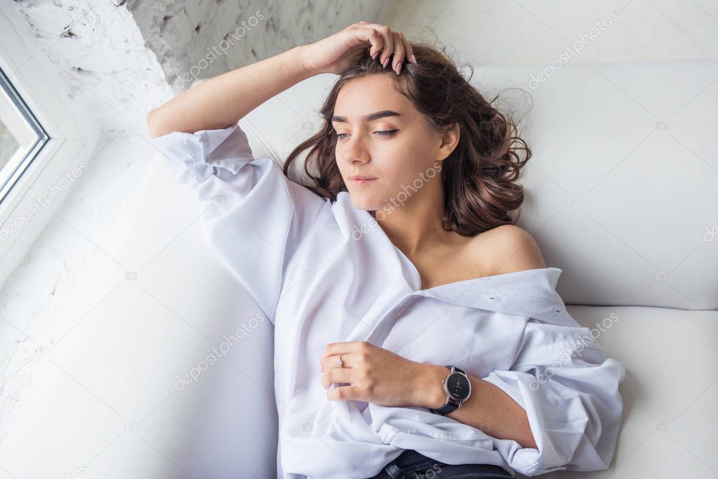young pretty sexy charming girl in a white man's shirt lying on a white sofa near the window in the bright room with her long hair loosened