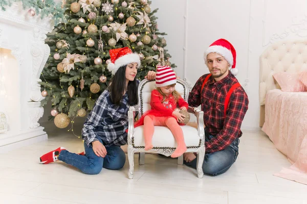 young father, mom\'s father dressed in festive clothing red hats, jeans and clashed shirts and a little girl in a red dress and hats sitting by the fir-trees and fireplace