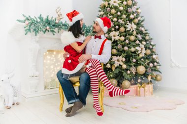 Sevimli, sevimli bir baştan çıkarıcı kız ve kocasının diz üzerine oturan kırmızı yılbaşı şapka içinde küçük bir sakallı bir adam bir Noel ağacı, şenlikli bir fotoğraf yakın: