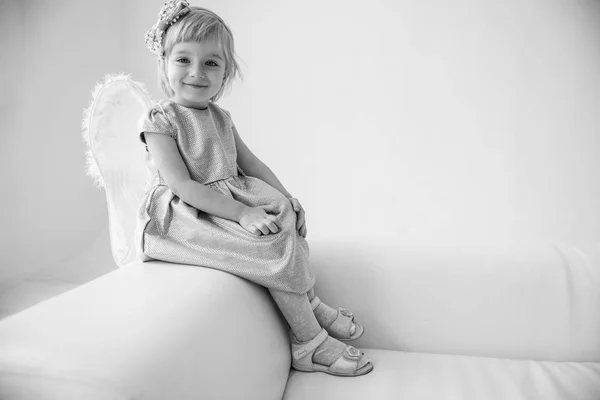 little cute little blonde girl in a gray dress with angelic white wings on a light sofa in the luminous room