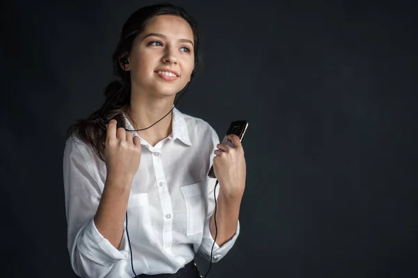 young pretty girl with brilliant hair with a nice smile, dressed in a white shirt on a gray background, holds small headphones for music and the phone and listens to the music